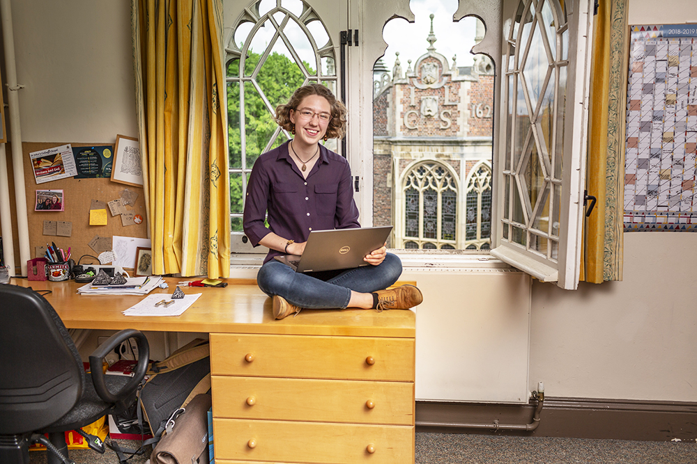 A student sits on the desk in her room with her laptop